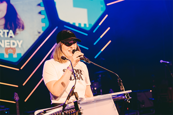 A woman speaks into a microphone on a colourfully lit stage, wearing a white t shirt and a baseball cap